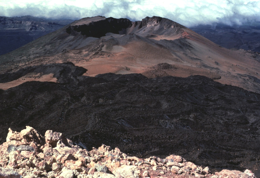 Fresh-looking lava flows, seen here from the summit of Pico de Teide, descend towards Pico Viejo (top center) and diverge to the north and south along a broad saddle between the two volcanoes.  A 750-m-wide crater truncates the summit of Pico Viejo, which was formed contemporaneously with Pico de Teide.  These two stratovolcanoes were constructed within the 10 x 17 km wide Las Cañadas caldera, whose floor is visible in the distance. Copyrighted photo by Katia and Maurice Krafft, 1977.