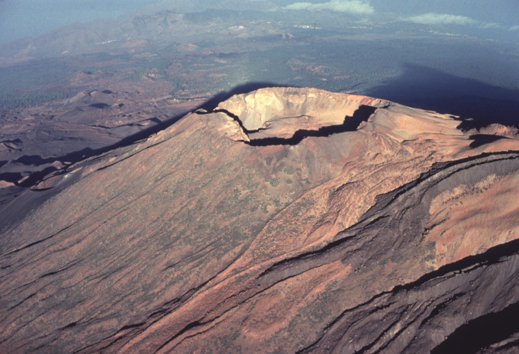 A prominent crater caps Pico Viejo, the second largest stratovolcano constructed within the massive Las Cañadas caldera.  The sharp-peaked summit of the largest post-caldera volcano, Pico de Teide, casts a shadow (upper right) on the floor of the caldera.  Dark-colored trachytic lava flows with prominent lateral levees descend at the lower right from the summit of Pico de Teide.  A chain of youthful cinder cones, some of which erupted during historical time, occupies the caldera floor beyond Pico Viejo at the left. Copyrighted photo by Katia and Maurice Krafft, 1977.