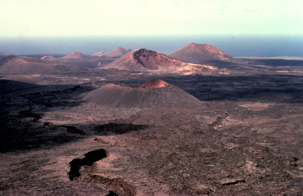 The largest historical eruption of the Canary Islands took place from 1730 to 1736, when a NE-SW-trending fissure formed the Montañas del Fuego ("Mountains of Fire").  Voluminous lava flows covering 200 km2 reached the western coast, seen in the distance, along a broad, 20-km-wide front.  The villages of Maretas and Santa Catalina were destroyed, along with the most fertile valleys and estates of the island, which was entirely evacuated.  Numerous cinder cones such as these dot the lava field, much of which lies within Timanfaya National Park.  Copyrighted photo by Katia and Maurice Krafft, 1977.