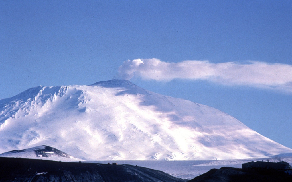 A gas plume from the Erebus summit crater casts a shadow on its western flank. There is an active lava lake in this crater and a caldera forms the plateau to the left of the summit cone. The McMurdo Station research facility on Hut Peninsula is in the foreground. Photo by Bill Rose, 1983 (Michigan Technological University).