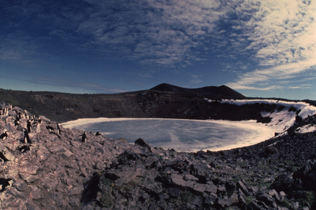 Petrel maar is a 300-m-wide crater on the eastern side of the roughly 1.5-km-wide Penguin Island.  The maar was formed during an explosive eruption in the early 20th century and is surrounded by angular blocks of ejecta.  The cinder cone behind Petrel maar to the SW is Deacon Peak, which marks the 180-m-high summit of the island.   Copyrighted photo by Katia and Maurice Krafft, 1984.