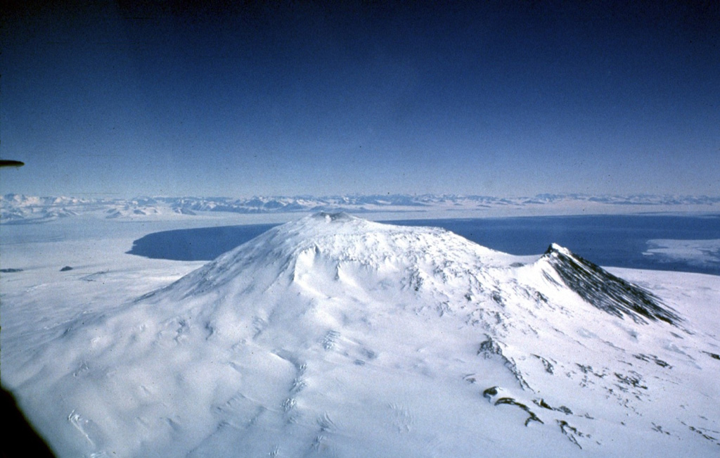 Mount Erebus is viewed here from the SE, with the water of the Ross Sea behind it and the mountains of Victoria Land in the distance. Fang Ridge is above the snow-free slope (right) on the NE flank. The plateau right of the summit is the lava flow-filled floor of the summit caldera, inside which the active summit cone was constructed. Hut Point Peninsula is the site of the McMurdo Station and is to the left of the Ross Sea. Photo by Bill Rose, 1983 (Michigan Technological University).