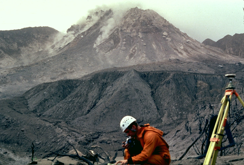 Scientists from the Montserrat Volcano Observatory make monitoring measurements in February 1997 as small rockfalls descend the flanks of the lava dome. Castle Peak lava dome, constructed during the previous eruption of Soufrière Hills during the 17th century, had collapsed three days before this photograph was taken from the Tar River Estate house, 2 km NE of the dome. Periodic collapse of the growing lava dome produced pyroclastic flows that in some cases reached the sea. Photo by Mark Davies, 1997 (Montserrat Volcano Observatory).