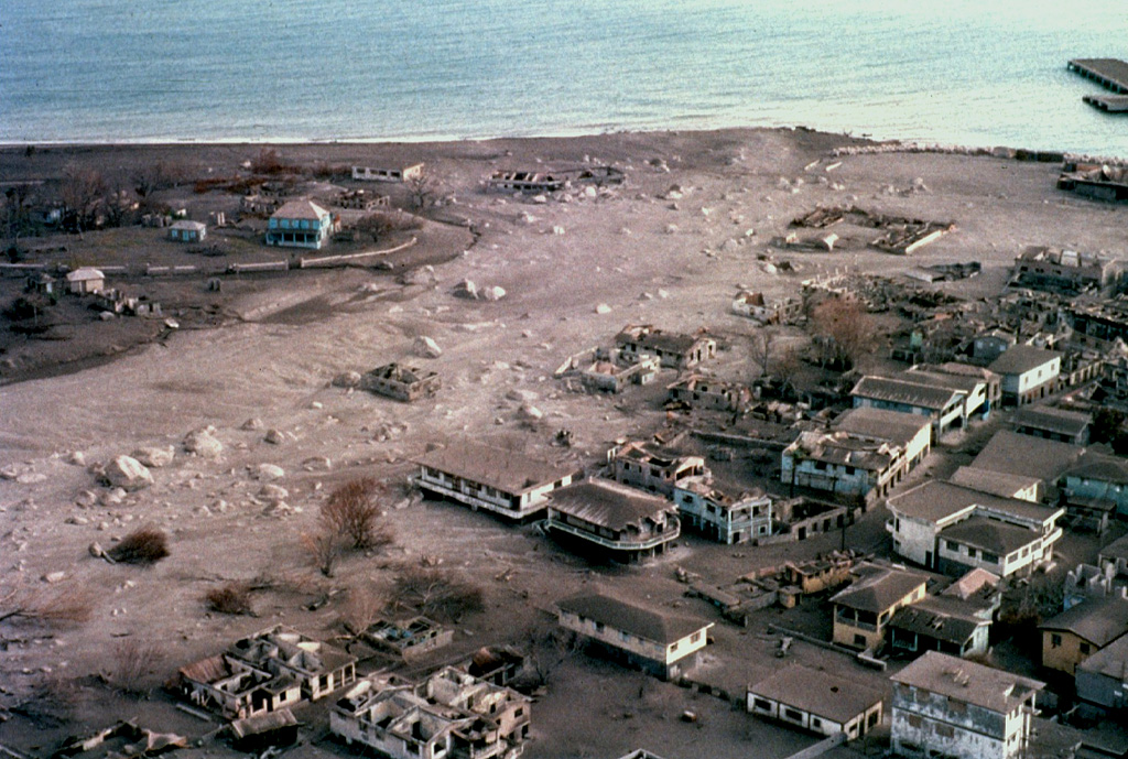 Pyroclastic flows and surges repeatedly swept through Plymouth, the former capital of Montserrat, beginning in August 1997.  This December 1997 photo shows the deserted city mantled by ash, with buildings partially or totally destroyed by the high-temperature pyroclastic flows.  Continued growth of the summit lava dome during 1997 had filled in the summit crater, which caused pyroclastic flows to spill over the western crater rim and travel down the Fort Ghaut valley into the evacuated city. Photo by Peter Francis, 1997 (Open University).