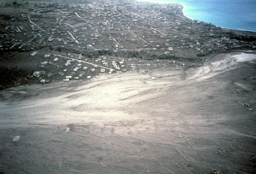 A broad plain of light-colored pyroclastic-flow deposits in the foreground partially buries the city of Plymouth, the former capital of Montserrat.  This December 1997 aerial view from the north shows Fort Ghaut valley, completely filled by pyroclastic-flow deposits, cutting diagonally across the center photo.  Sugar Bay lies at the upper right, just south of the port of Plymouth.  Pyroclastic flows began entering Plymouth in August 1997. Photo by Mark Davies, 1997 (Montserrat Volcano Observatory).