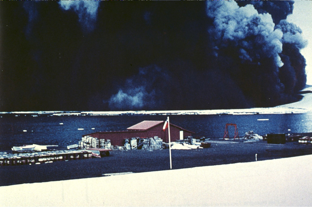 A wide, dark, ash-laden eruption plume towers above Pendulum Bay on 4 December 1967, originating from vents located about 2 km NW of the Chilean research station seen in the foreground. Heavy ashfall from the eruption severely damaged the Chilean base. The destruction of the base was completed by a subsequent eruption in 1969. Members of the research party walked about 6 km S to safety at the British research station on the first day of the eruption and were then evacuated by helicopter. Photo by Bernardo Blass, 1967 (published in González-Ferrán, 1995).