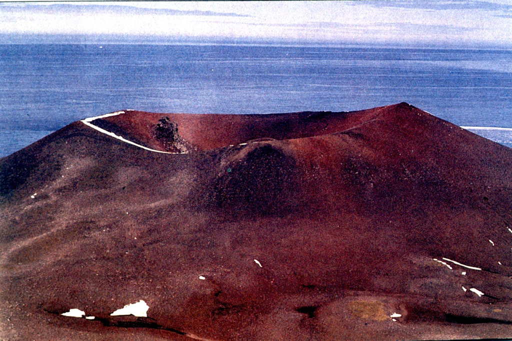 Deacon Peak scoria cone on Penguin Island contains a summit crater approximately 350 m wide and 75 m deep. The upper slopes of the cone are composed of reddish oxidized pyroclastic rocks.  The formation of Deacon Peak was dated to about 1679 CE using lichenometry, which is based on the calibration of lichen growth rates. Photo by Oscar González-Ferrán (University of Chile).