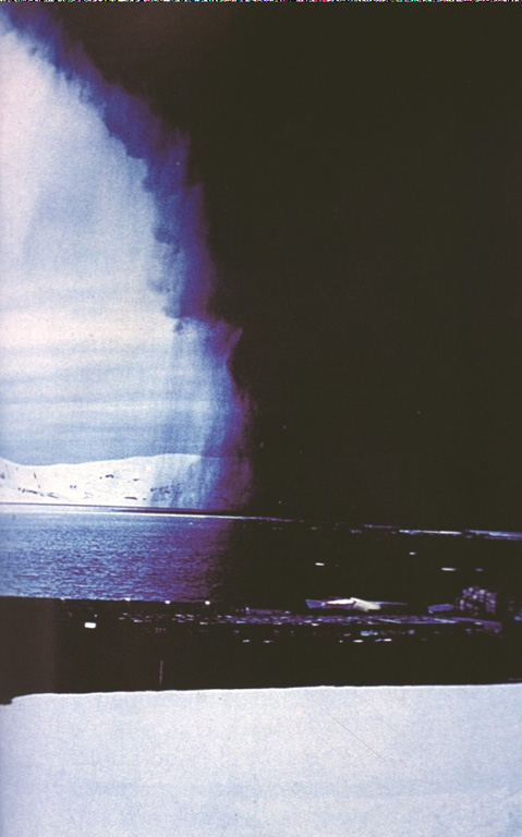 An eruption darkens the sky above Antarctica's Deception Island in December 1967. Falling ash can be seen trailing from the margins of the eruption cloud. Winds distributed the ash plume to the NE and deposited 30 cm of ash over a 2 km swath across Port Foster caldera bay onto the Chilean Antarctic research station, where this photo was taken. Photo by Bernardo Blass, 1967 (published in González-Ferrán, 1995).