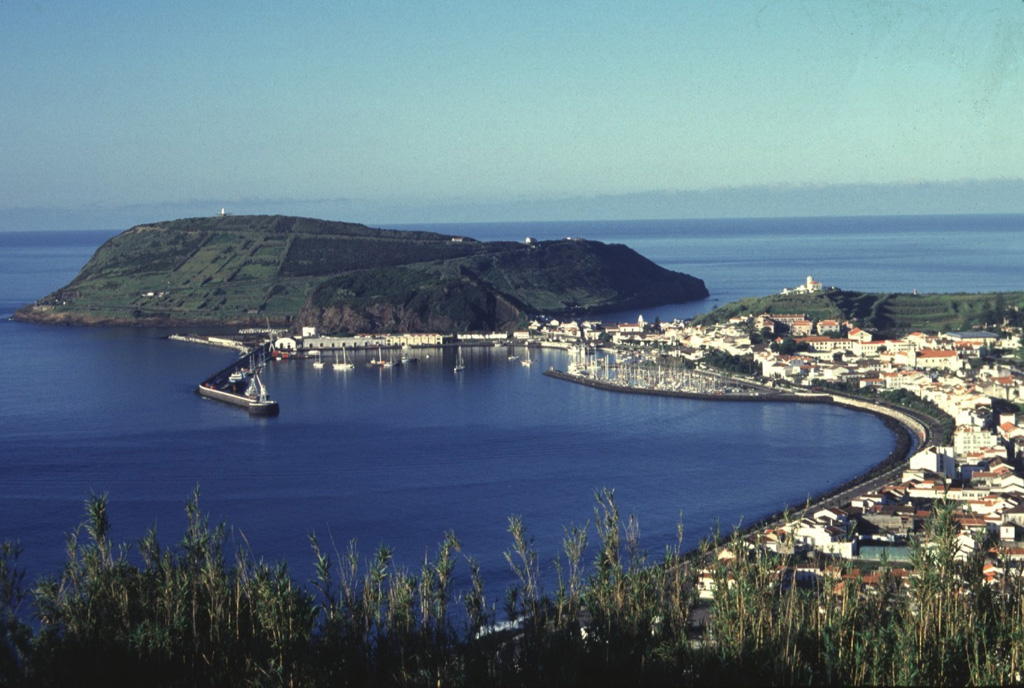 The Monte da Guia tuff cone forms a backdrop to the harbor of Horta, the largest town on the island of Fayal. The cone was formed by phreatomagmatic eruptions just off the SE tip of the island and is now connected to Fayal by a low narrow peninsula. It is one of a cluster of cones on the lower SE flank. These cones and an associated basaltic lava field form the late Pleistocene Almoxarife Formation. Photo by Rick Wunderman, 1997 (Smithsonian Institution).