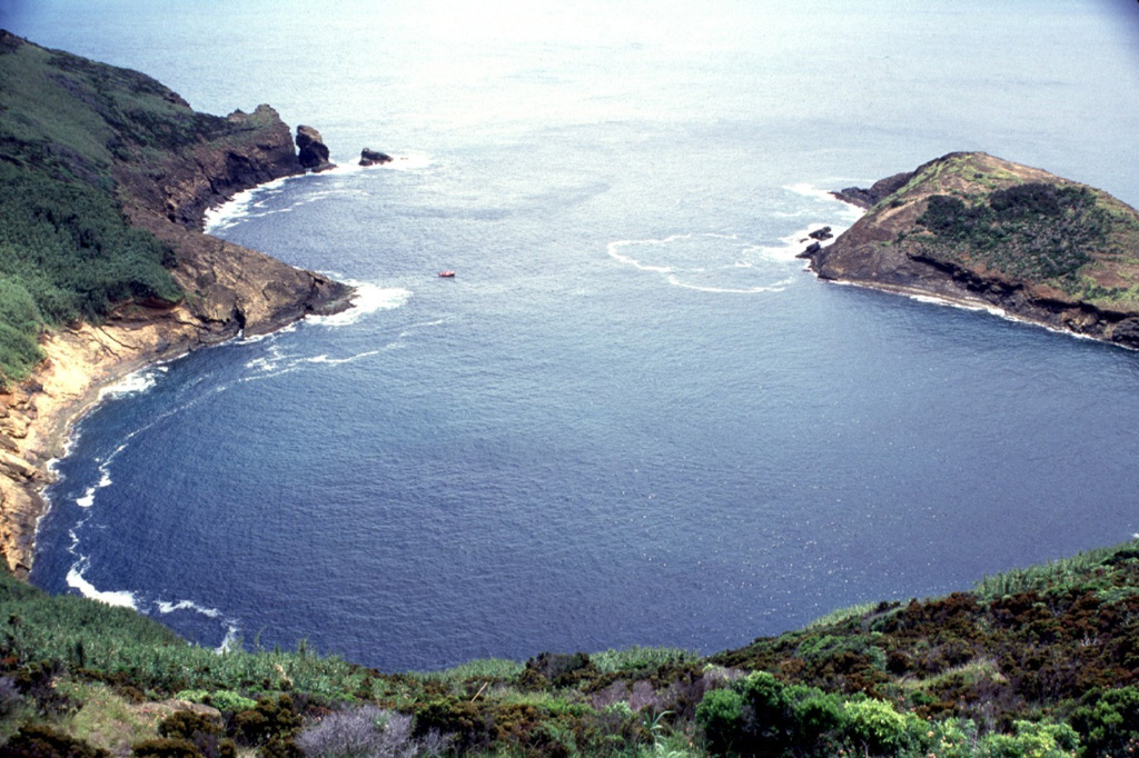 The crater of Monte da Guia tuff cone at the SE tip of Fayal Island has been breached by the sea, forming a circular bay measuring about 500 m long. This view looks out to sea from the NW rim of the cone, which was formed by phreatomagmatic eruptions produced when magma encountered water along the coast. A small boat used by divers provides scale off the headland at left center. Photo by Rick Wunderman, 1997 (Smithsonian Institution).