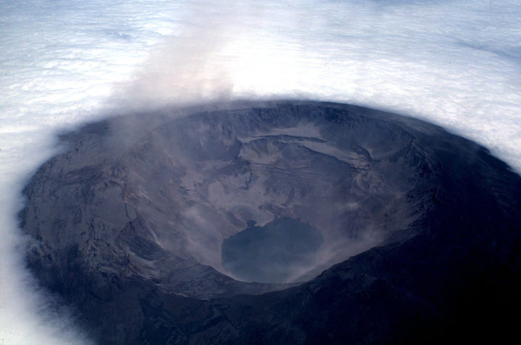 Dust clouds rise from Fernandina caldera on July 4, 1968, about three weeks after a major explosive eruption that was followed by collapse of the caldera floor.  Collapse occurred incrementally and asymmetrically, ranging up to about 350 m at the SE end of the caldera, which contains the caldera lake.  Fernandina, the most active of Galápagos volcanoes, is a basaltic shield volcano with a deep 4 x 6.5 km summit caldera.  Flat-topped benches mark the SE and NW ends of the elliptical caldera. Photo by Tom Simkin, 1968 (Smithsonian Institution).