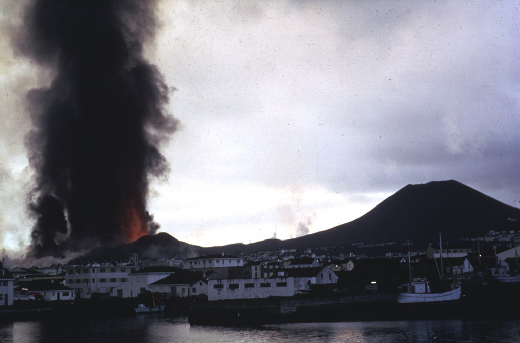 A dark ash column rises above lava fountains from a fissure eruption on the island of Heimaey in 1973. This explosive activity produced the Eldfell cinder cone (left), followed by effusion of lava flows which threatened the town of Vestmannaeyjar (foreground). Helgafell cone, on the right, formed during an eruption 5,900 years ago. Photo by Tom Simkin, 1973 (Smithsonian Institution).