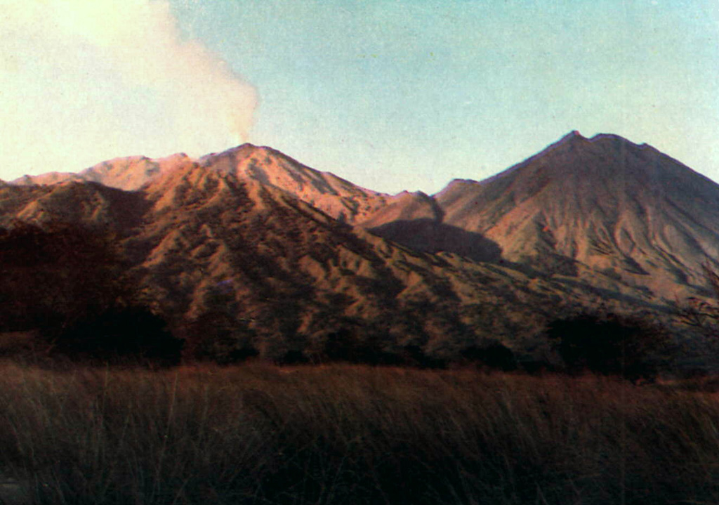A plume rises above Doro Api, one of the two large post-caldera cones of Sangeang Api, in 1985. Doro Mantai appears at the right in this view from Sori Buntu on the S flank. An eruption that included both explosive activity and the effusion of lava flows down the W flank began in July 1985 and lasted until February 1988. Photo by Samud W., 1990 (Volcanological Survey of Indonesia).