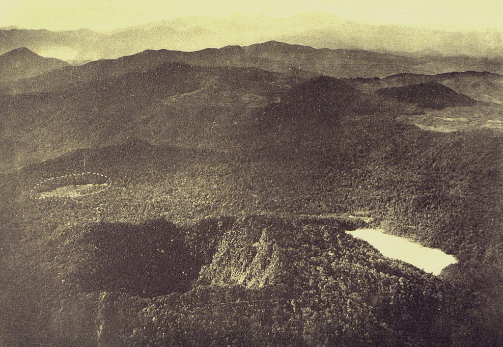 An aerial view from the NNW shows the two summit craters of Gunung Patuha volcano in the foreground. Steep-walled Taman Saat crater (left) is located NW of lake-filled Kawah Putih (right). The dashed circle at the middle left indicates an unvegetated area called the “Tugeslibde crater.” Photo published in Taverne, 1926 