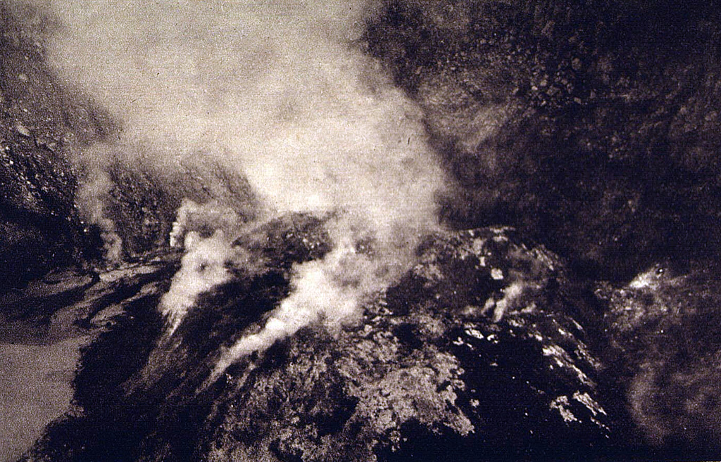 A plume rises above a lava dome emplaced in the summit crater of Lewotobi Perempuan volcano in 1921. The 90-130 m wide, 35-m-high lava dome is seen here from the NW crater rim. Photo published in Kemmerling 1929, 