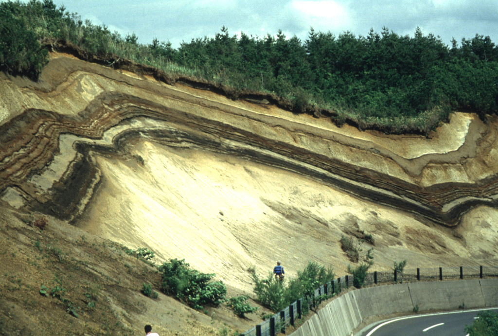 A roadcut west of the Ninokura dam exposes deposits from Towada. The thick light-colored unit at the base is the Hachinohe ignimbrite that formed about 13,000 years ago during the last of a series of explosive eruptions that resulted in the incremental formation of Towada's 11-km-wide caldera. The thinner light- and dark-colored deposits above the Hachinohe ignimbrite were produced by post-caldera eruptions, the most recent of which took place in 915 CE. Photo by Yukio Hayakawa (Gunma University).