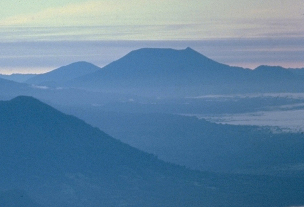 The truncated summit of Gunung Ibu stratovolcano along the NW coast of Halmahera is seen here from the SW at the summit of Gamalama volcano.  The broad summit of Gunung Ibu has nested summit craters.  The inner crater, 1 km wide and 400 m deep, contains several small crater lakes.  Only two eruptions are known from Ibu in historical time, a small explosive eruption from the summit crater in 1911, and another beginning in December 1998.     Copyrighted photo by Katia and Maurice Krafft, 1976.