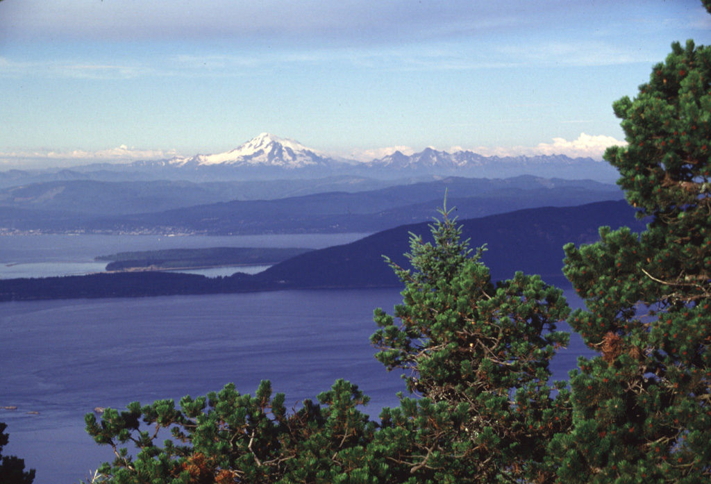 Mount Baker forms a prominent landmark visible throughout much of the northern Puget Sound region. Its glaciated slopes rise above Bellingham Bay, as seen here from Mount Constitution on Orcas Island. Lummi Island, another of the San Juan Islands, forms the ridge extending across the center of the photo. The Twin Sisters massif, composed of olivine-rich ultramafic rocks derived from the Earth's mantle, is located immediately SW (right) of Mount Baker. Photo by Lee Siebert, 1998 (Smithsonian Institution).