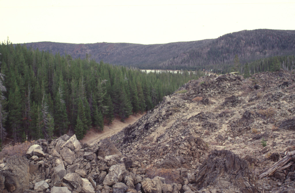 The East Lake obsidian flows and associated pumice deposits erupted from fissures parallel to the inner caldera wall. The westernmost of the two obsidian flows immediately south of East Lake is seen here. It traveled to the north towards East Lake, part of which can be seen beyond the forested area in the center of the photo, below the eastern caldera wall.  Photo by Lee Siebert, 1997 (Smithsonian Institution).