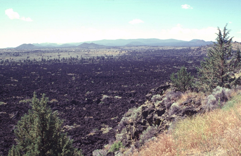 Despite its youthful appearance, the Devils Homestead lava flow in the foreground is slightly more than 10,000 years old.  Erosional modification of lava flow surfaces proceeds slowly in arid regions.  The Devils Homestead flow is one of the largest on the north flank of Medicine Lake volcano, the broad shield volcano on the horizon.  Flank cinder cones dot the slopes of the chemically diverse shield volcano, which has produced both the basaltic cinder cones and lava flows seen here, and dacitic and rhyolitic pumice and lava flows. Photo by Lee Siebert, 1998 (Smithsonian Institution).