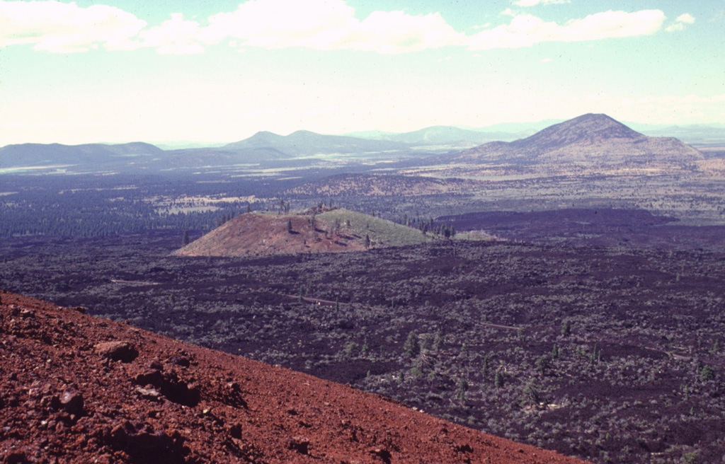 The basaltic Callahan lava flow, erupted from the Cinder Butte cone, is the largest Holocene lava flow on the north flank of Medicine Lake volcano.  The broad Callahan flow is seen here from the reddish oxidized rim of Cinder Butte diverging around an older cinder cone to the north.  The flow was dated from radiometric and paleomagnetic evidence at about 800 CE.  The Callahan flow is chemically zone, with initial lavas being andesitic and later lavas basaltic. Photo by Lee Siebert, 1998 (Smithsonian Institution).