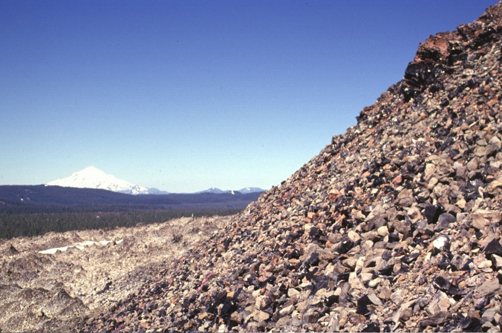 Blocks of glassy obsidian reflect sunlight from the surface of a rhyolitic lava dome marking the latest eruptive event during extrusion of the Glass Mountain lava flow.  The composite flow originated from more than a dozen NW-SE-trending vents near the eastern rim of the Medicine Lake caldera.  Part of the massive obsidian flow seen on the left side of the photo traveled west into the caldera.  Snow-capped Mount Shasta towers above the forested caldera rim to the west. Photo by Lee Siebert, 1998 (Smithsonian Institution).