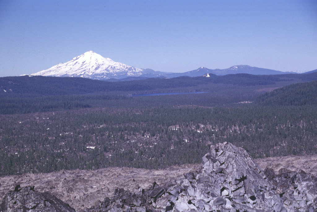 The broad partially forested lava flow extending across the center of the photo into Medicine Lake caldera is the Hoffman lava flow, which erupted about 1,170 years ago. The massive rhyolite lava flow and an adjacent smaller one were erupted from vents on the NE rim of the caldera. The larger of the two flows, seen here with the unvegetated Glass Mountain flow in the foreground, was erupted near the caldera rim and flowed both west into the caldera and down the east flank. Snow-capped Mount Shasta rises to the west. Photo by Lee Siebert, 1998 (Smithsonian Institution).