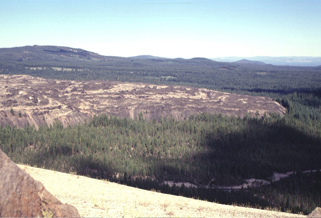The steep-sided SW margin of the Little Glass Mountain obsidian flow rises above the surrounding forested flanks of Medicine Lake volcano. Eruption of the rhyolitic lava flow was preceded by a pumice eruption that mantled the surrounding area (including the basaltic cone of Pumice Stone Mountain in the foreground) with white pumice. The 0.4 km3 Little Glass Mountain lava flow is the second largest silicic lava flow at Medicine Lake. Photo by Lee Siebert, 1998 (Smithsonian Institution).