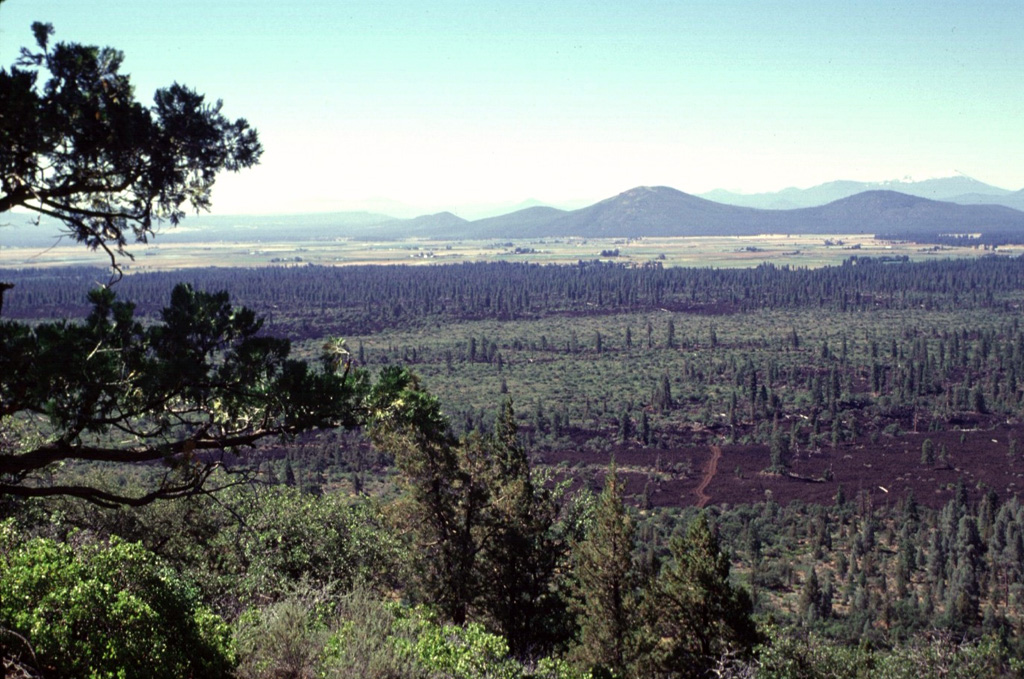 The sparsely vegetated lava flows in the foreground and the darker-colored forested flows in the middle of the photo were erupted from Brushy Butte, a low shield volcano capped by a cinder cone.  The flows extend down the Falls River valley to the south.  The two larger peaks in the right background, located south of the town of Fall River Mills, are Bald Mountain and Cinder Butte. Photo by Lee Siebert, 1998 (Smithsonian Institution).