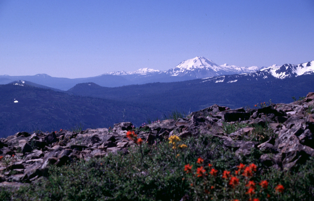 Eiler Butte, the small flat-topped cinder cone at left-center just below the low point on the horizon, is the northermost of a chain of cinder cones forming the Tumble Buttes volcanic field.  Eiler Butte, located within the Thousand Lake Wilderness Area, is seen here from Burney Mountain, with snow-capped Lassen Peak in the background to the south.  The Tumble Buttes are a N-S-trending chain of cinder cones and lava flows that form some of the youngest volcanic features north of Mount Lassen. Photo by Lee Siebert, 1998 (Smithsonian Institution).