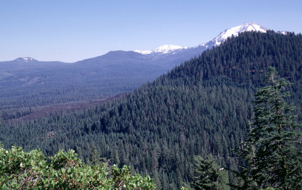 Bear Wallow Butte, the forested peak at the right, was the source of the youthful-looking unvegetated lava flows seen at the left-center.  Bear Wallow Butte lies at the southern end of a N-S-trending chain of vents known as Tumble Buttes.  Snow-capped Lassen Peak is visible to the south just beyond the summit of Bear Wallow Butte. Photo by Lee Siebert, 1998 (Smithsonian Institution).