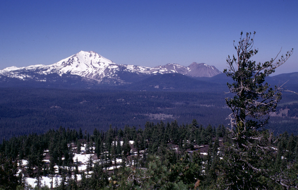 Snow-capped Lassen Peak is seen here from the summit of Prospect Peak shield volcano at the NE end of Lassen Volcanic National Park.  The unvegetated, largely snow-free peaks on the right horizon are the Chaos Crags, a complex of dacitic lava domes last active about 1100 years ago.  The Twin Lakes sequence of andesitic lava shields and cones forms the lake-studded area of the Central Plateau in the center of the photo. Photo by Lee Siebert, 1998 (Smithsonian Institution).