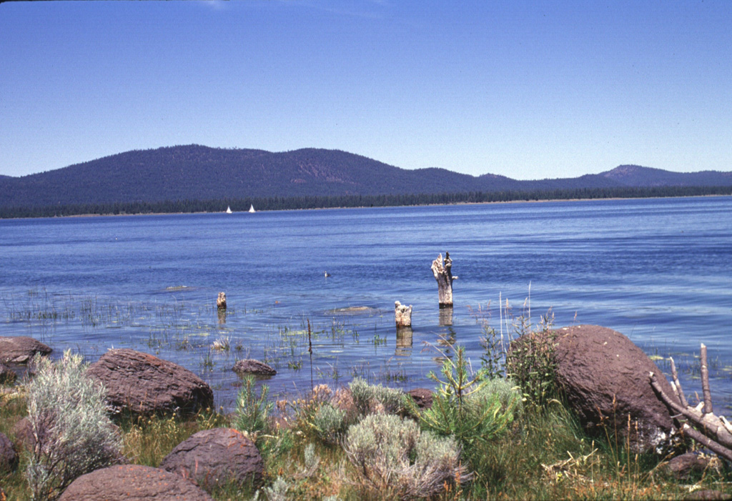 Forested cinder cones of the Eagle Lake volcanic field rise above the SW shore of Eagle Lake.  The volcanic field occupies the junction of the Cascades, Sierra Nevada, and Basin and Range geologic provinces.  The Eagle Lake field consists of 15 cinder cones and basaltic lava flow vents within a larger Quaternary basaltic field.  The vents are aligned along faults defining the Eagle Lake volcano-tectonic depression.  The latest eruptive period occurred during the late-Pleistocene or early Holocene. Photo by Lee Siebert, 1998 (Smithsonian Institution).