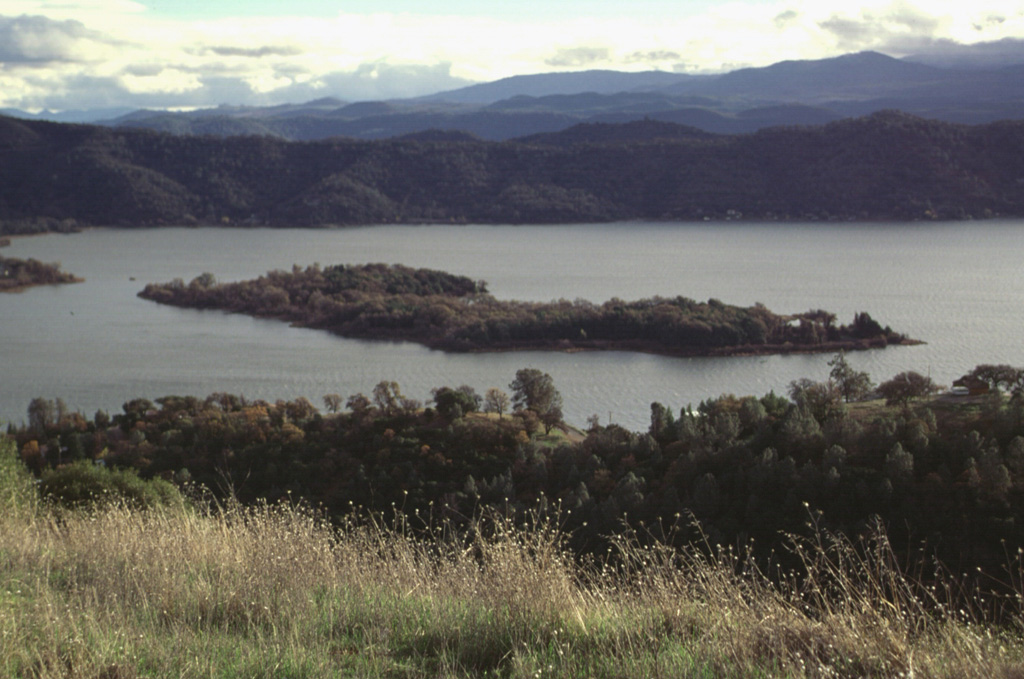 Elongated Rattlesnake Island near the NE tip of Clear Lake is seen here from the NW.  Most of the flat-lying western side of the island is a basaltic-andesite lava flow of late-Pleistocene age.  The flow was erupted to the NE and NW from a cinder cone that is the gently rounded ridge that forms the far southern tip of the island at the left.  The scoriaceous aa flow was erupted subaerially above the lake level. Photo by Lee Siebert, 1997 (Smithsonian Institution).