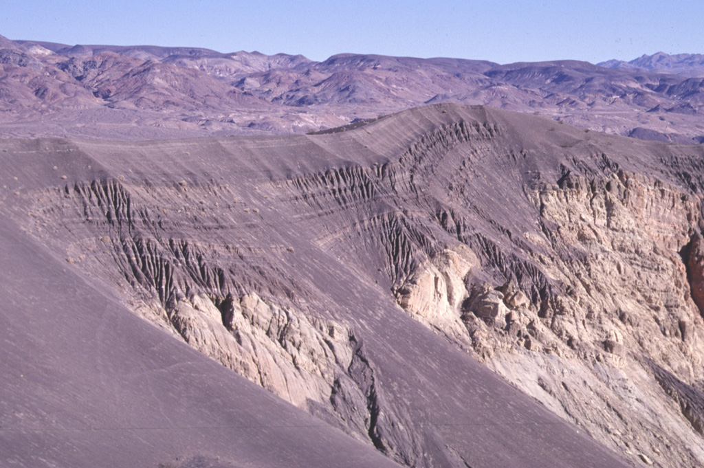 Darker-colored layers of basaltic ash drape the northern rim of Ubehebe Crater in the northern part of Death Valley National Park and spill down the crater walls.  The 235-m-deep Ubehebe Crater is the largest of a series of more than a dozen overlapping maars formed by explosive eruptions through fanglomerate deposits, which form the light-colored areas below the ash layers. The Amargosa Range rises on the horizon across Death Valley to the east. Photo by Paul Kimberly, 1997 (Smithsonian Institution).