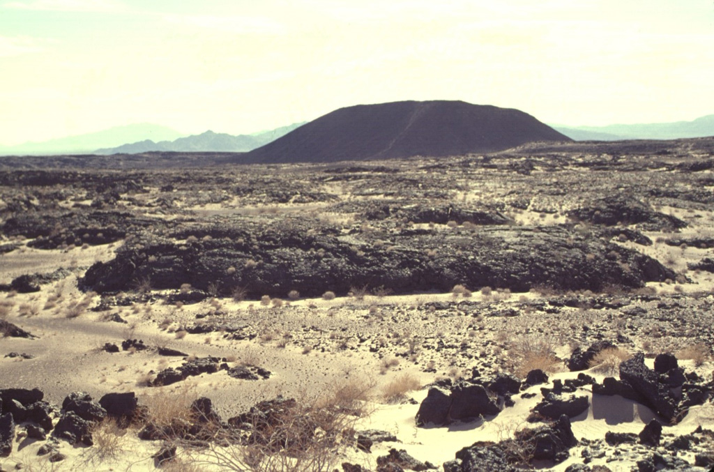 Amboy Crater is an isolated 75-m-high cinder cone seen here from the NW with the Bullion Mountains in the background.  Amboy was the source of a 70-sq-km basaltic lava field that was erupted during the late Pleistocene onto a flat-lying alluvial valley.  The dominantly pahoehoe lava field divided the Bristol Dry Lake into two playas.  Depressions on the surface of the lava flow are filled with light-colored windblown sand distributed by prevailing winds from the west.  The light-colored diagonal stripe on the flank of the cone is a trail leading to the crater rim.  Photo by Lee Siebert, 1997 (Smithsonian Institution).