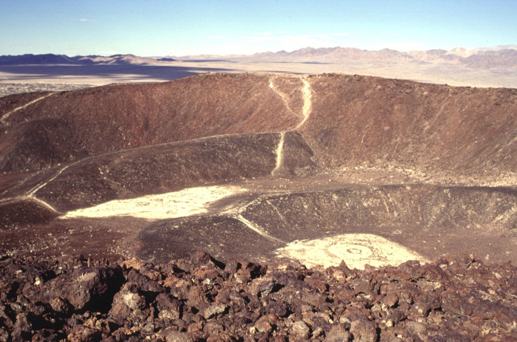 Amboy Crater is a complex cone formed of at least four nested craters.  The rims of two relatively undissected inner cones can be seen in this view from the SE rim of the outer crater of the 75-m-high cinder cone.  The two outer cones are breached on their western sides, out of view to the left.  Amboy crater is surrounded by a 70-sq-km basaltic lava field.  The Bristol Mountains form the skyline. Photo by Lee Siebert, 1997 (Smithsonian Institution).