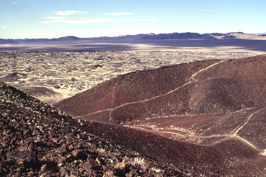 Amboy Crater is a composite cinder cone that contains at least four nested craters.  The two outermost craters are breached to the west.  A broad lava field that extends from the cone is visible beyond the breach.  Light-colored windblown sand deposited by prevailing winds from the west fill in low spots on the surface of the lava flow. Photo by Lee Siebert, 1997 (Smithsonian Institution).