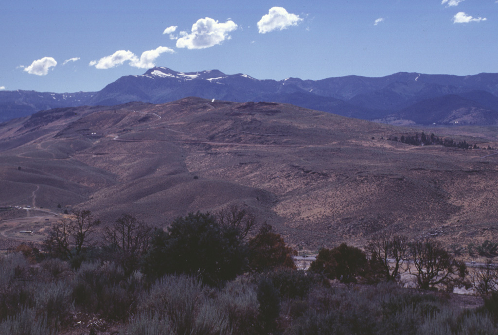 Steam rises from a geothermal well at the top of a lava-dome complex of the Steamboat Hills geothermal field in west-central Nevada south of Reno.  U.S. Highway 395 traverses the valley beyond the trees in the foreground.  This is the site of the Steamboat Resort, which dates back to the 1860s, when it was frequented by miners working on the nearby renowned Comstock Lode.  The Carson Range forms the ridge on the horizon. Photo by Lee Siebert, 1998 (Smithsonian Institution).