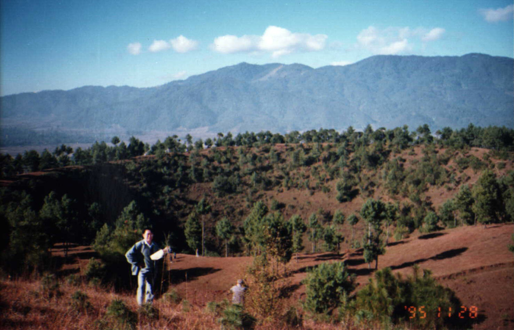 A geologist stands at the rim of a crater in the Tengchong Volcanic Field, with Ailuo Mountain in the background to the W. The youngest eruptions from the Tengchong field, which surrounds the city of Tengchong, produced olivine basalts and basaltic andesites. Photo by Liu Xiang, 1995 (Changchun University).