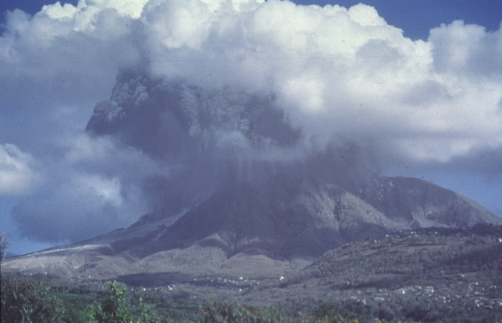 A dark ash column rises into light-colored weather clouds surrounding Soufrière Hills volcano.  This photo was taken on October 20, 1997 from the old volcano observatory 7 km NW of the summit about a minute after the onset of the explosion.  Partial collapse of the eruption column soon produced pyroclastic flows that descended the eastern and western flanks.  During the course of the eruption that began in 1995 the volcano observatory had to be moved farther away from the volcano several times as the risk from eruptions increased. Photo by Paul Cole, 1997 (Montserrat Volcano Observatory).