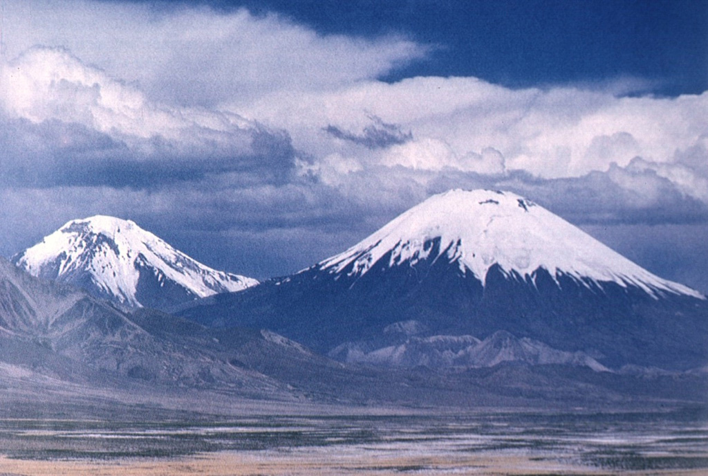 The Nevados de Payachata volcanic group in northern Chile, seen here from the west, consists of the symmetrical, 6348-m-high Parinacota volcano (right) and its older twin volcano, Pleistocene 6222-m-high Pomerape volcano (left).  The young cone of Parinacota post-dates collapse of an older edifice about 8000 years ago.  The most recent activity at Parinacota produced a series of fresh-looking lava flows from satellitic cones on the south and SW flanks.   Photo by Oscar González-Ferrán (University of Chile).
