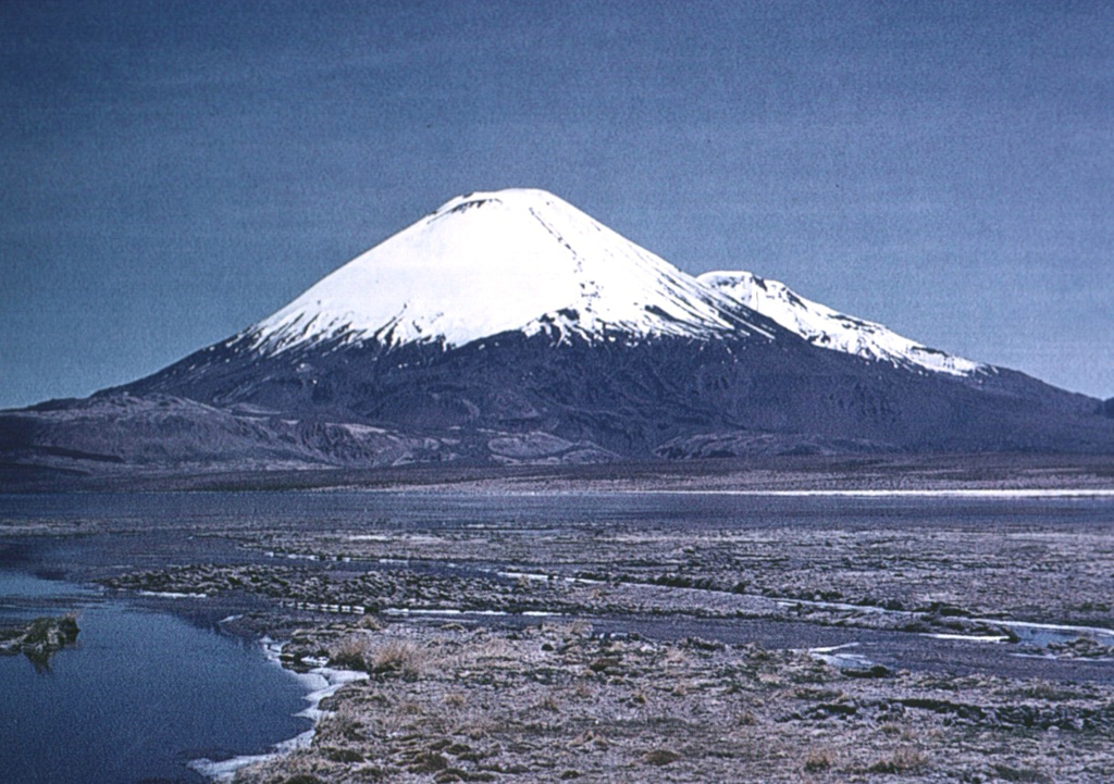 The southern side of conical, glacier-clad Parinacota volcano is seen from south of Laguna Changará, with its twin volcano, Pomerape, visible in the distance behind its right-hand flank. A complex of lighter colored dacitic-rhyolitic lava domes can be seen at the SW flank of Parinacota (middle left).  The main cone of Parinacota was constructed during the Holocene primarily by the effusion of andesitic lava flows following collapse of an earlier edifice.  The youngest of these flows was dated at between 1300 and 2000 years ago. Photo by Oscar González-Ferrán (University of Chile).