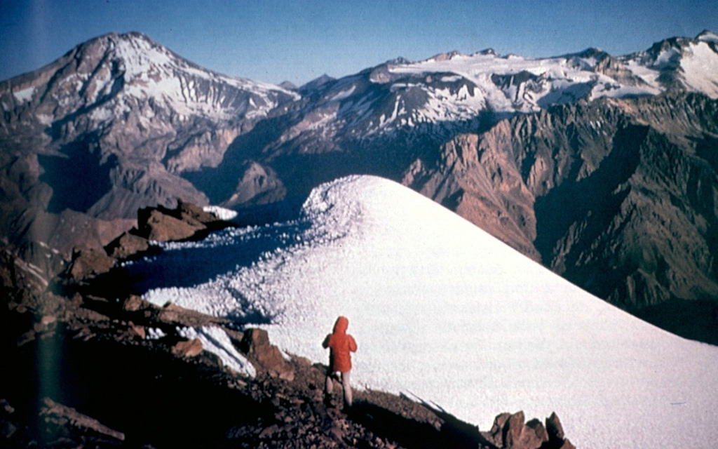 Tupungatito volcano, the northernmost historically active volcano of the central Chilean Andes, is the broad, glacier-clad massif at the right center.  The glacial icecap fills the southern side of the Pleistocene Nevado Sin Nombre caldera, which is breached to the NW, in the direction of this photo.  A dozen Holocene craters are found at Tupungatito, which has produced frequent mild explosive eruptions during the 19th and 20th centuries.  Tupungatito is located immediately SW of Pleistocene Tupungato volcano, the large conical peak at the left.   Photo by Sergio Kunstmann-Z, courtesy of Oscar González-Ferrán (University of Chile).