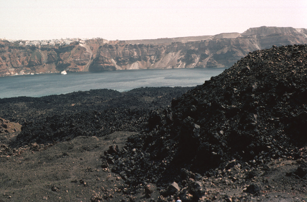The black lava flows that descend into Santorini's caldera bay are the Dafni lavas, erupted during 1925-26. During this eruption, which began in August 1925 and lasted until January 1926, Nea Kameni became a single island as the Dafni lavas united Mikra Kameni, Nea Kameni, and the Georgios domes. Eruptions resumed in May 1926, producing small pyroclastic flows. The steep-sided eastern caldera wall rises in the distance, capped by the town of Fira and the smooth-textured pyroclastic-flow deposits of the Minoan eruption. Photo by Lee Siebert, 1994 (Smithsonian Institution)