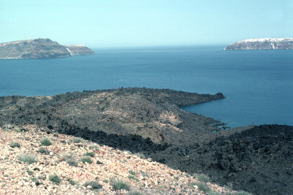 The lava flow forming the peninsula (center) dates back to an eruption during 1707-1711. Nea Kameni Island was formed between Palaea and Mikri Kameni islands during alternating explosive and effusive activity that began with uplift of a small islet called Aspronisi (White Island). The islands of Thirasia (left) and Thira (right) along the caldera rim can be seen in the distance to the NW. Photo by Lee Siebert, 1994 (Smithsonian Institution)