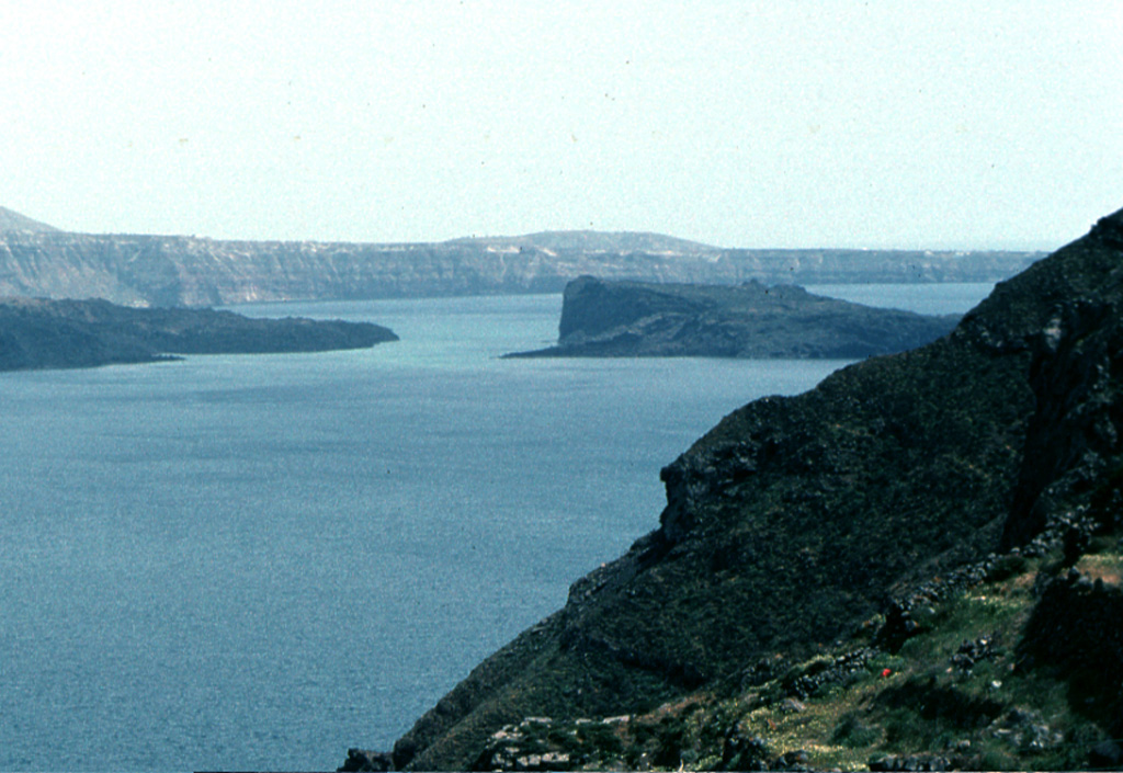 Palaea Kameni Island (right-center) rises from Santorini's caldera bay SE of the foreground cliffs of Thirasia Island. The higher, flat-topped part of the island was formed during 46-47 CE, and the slopes in front of it date back to 726 CE. The highly explosive eruption of the summer of 726 produced great quantities of pumice and ash that were transported as far as the islands of Abydos and Lesbos, Asia Minor, and Macedonia. Extrusion of the Ayios Nikolaos lavas followed, which eventually joined the NE tip of Thia (Palaea Kameni) Island. Photo by Lee Siebert, 1994 (Smithsonian Institution)
