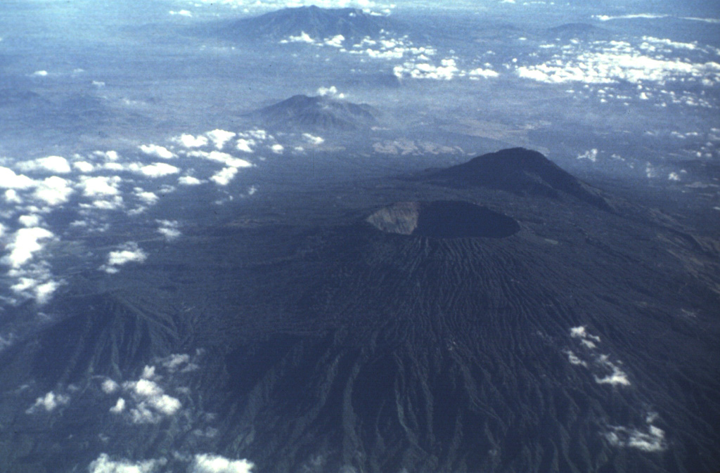 Boquerón volcano, its summit cut by a steep-walled, 500-m-deep crater, was constructed within a 6-km-wide caldera whose largely obscured rims are visible in this aerial photo from the WSW.  The caldera cut an older San Salvador edifice, remnants of which are visible at El Picacho peak (in the shadow behind Boquerón) and El Jabalí (the low peak at the lower left).  The buried caldera rim in the foreground is defined by the change in degree of dissection of the volcano's flanks.  Guazapa volcano can be seen in the distance at the top of the photo. Photo by Carlos Pullinger, 1996 (Servicio Nacional de Estudios Territoriales, El Salvador).