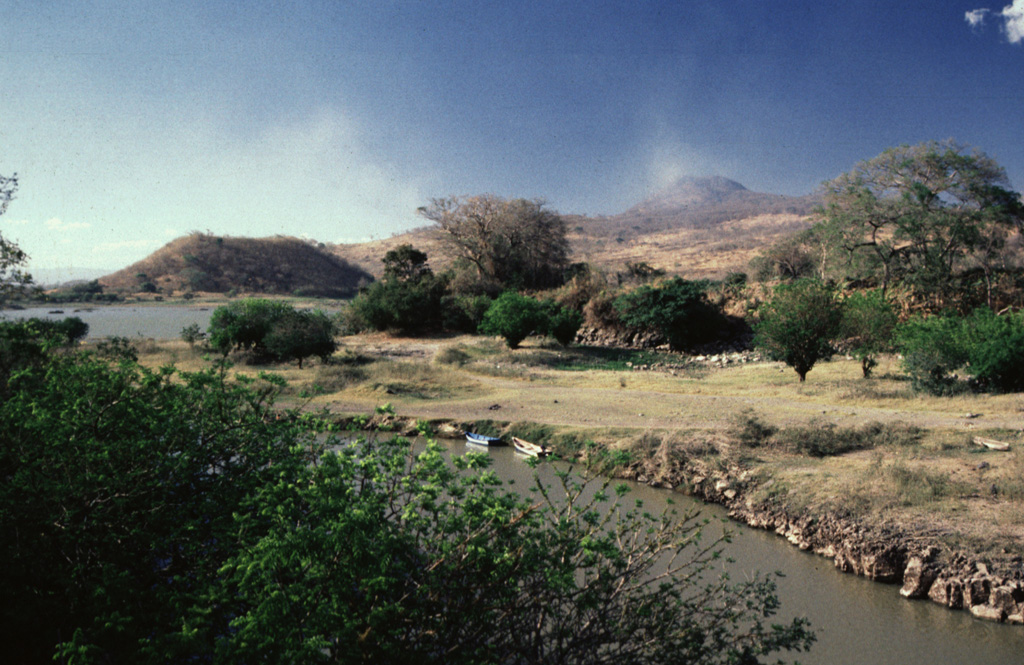Volcán de San Diego (upper right) is seen here from the south at the outlet of Lake Güija. The lake formed after lava flows from San Diego blocked the channel of the Desagüe river. Cerro el Tule (left) lies across a narrow channel from the lower flanks of San Diego. Cerro el Tule contains a summit crater and is one of the many cones of the San Diego volcanic field.  Photo by Giuseppina Kysar, 1999 (Smithsonian Institution).