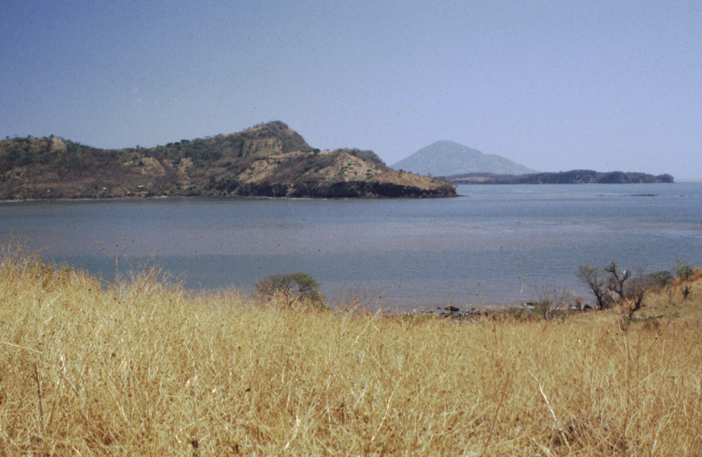 Isla Zacatillo lies in the Gulf of Fonseca only 2 km from Punta el Chiquirín, the eastern tip of El Salvador.  The low, roughly 3-km-wide island is crudely rectangular in shape, but has a peninsula extending to the NE.  A steep escarpment truncates the western side of the island, seen here.  NNW-SSE-trending faults cut lava flows on the island.  The conical peak in the distance is Isla el Tigre in Honduras. Photo by Rick Wunderman, 1999 (Smithsonian Institution).